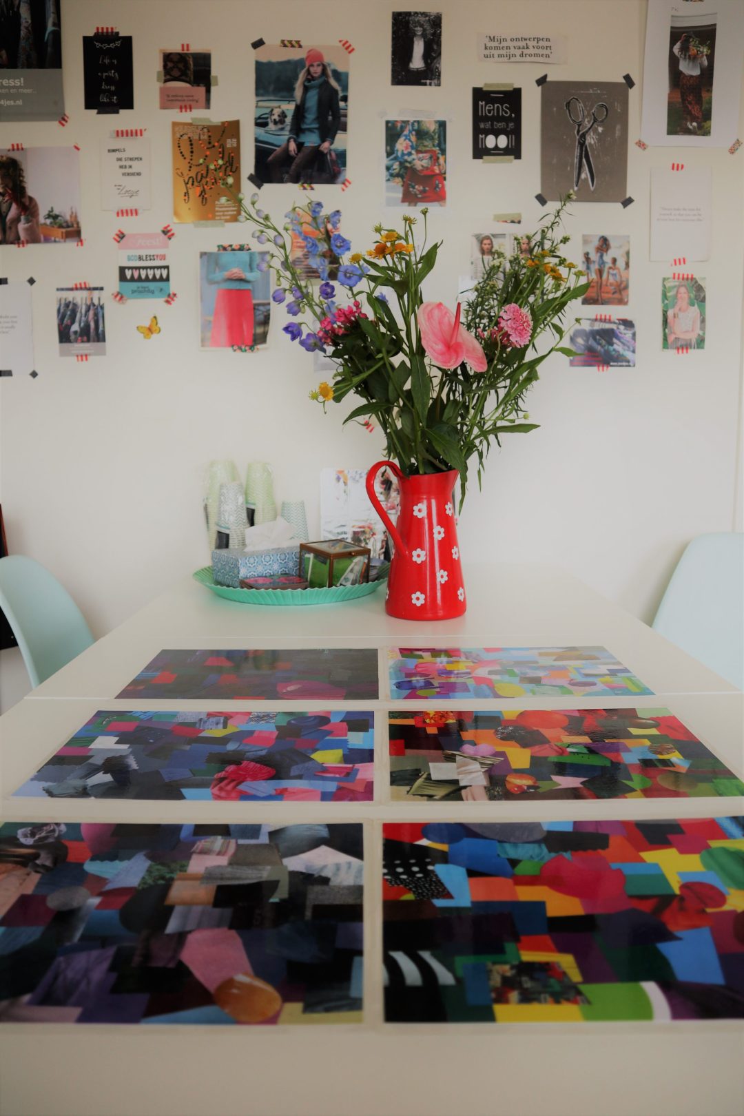Studio met kleurcollages op tafel, bloemen in een rode kan en een inspiratiewand