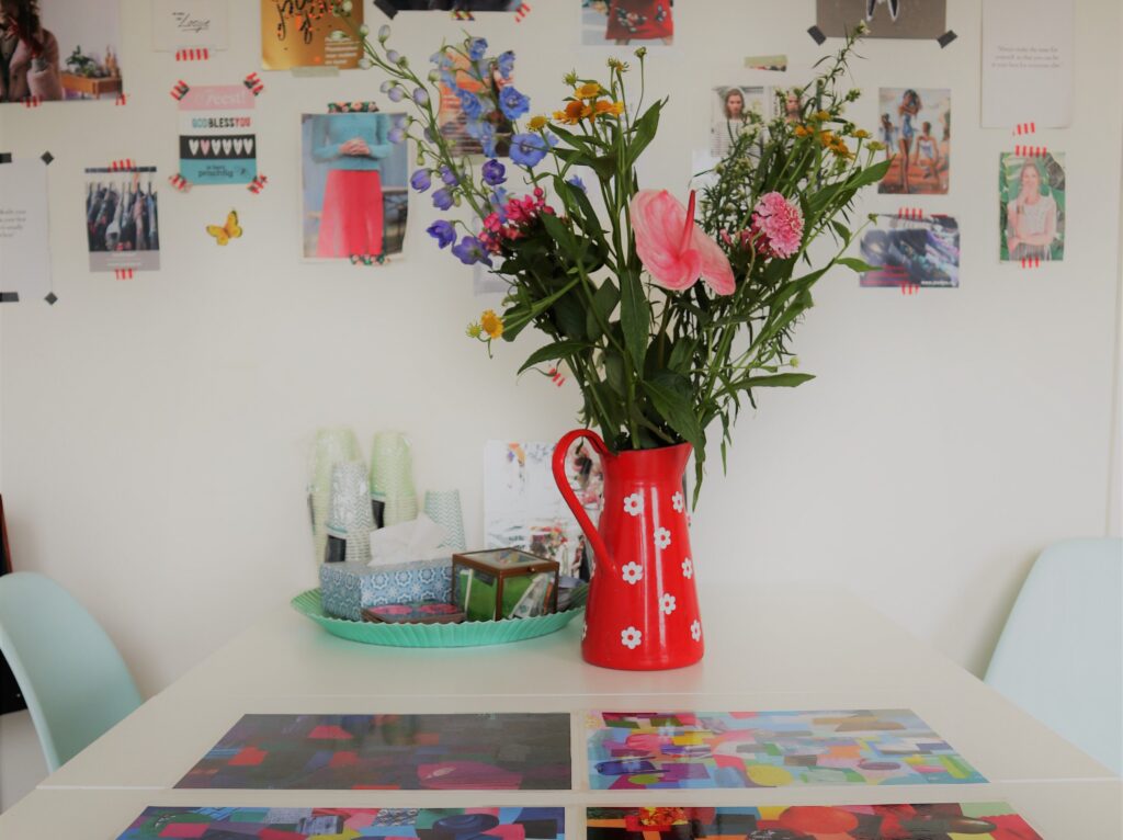 Studio met kleurcollages op tafel, bloemen in een rode kan en een inspiratiewand